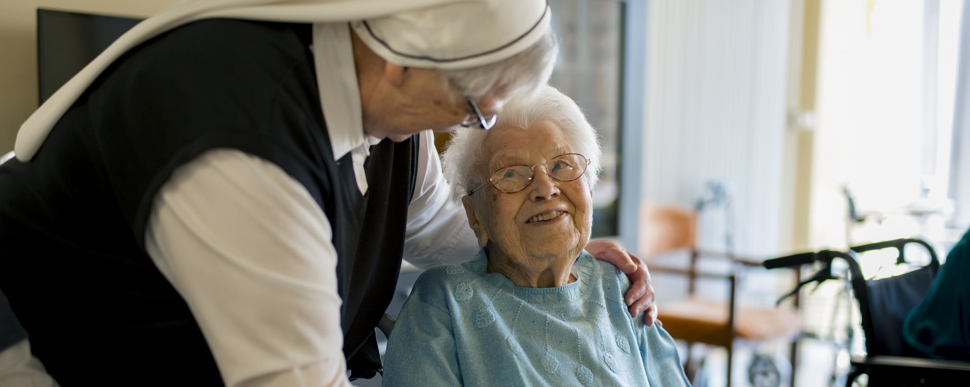 Eine Ordensschwester steht neben einer Frau, die im Rollstuhl sitzt und hat ihr einen Arm um die Schultern gelegt, mit der anderen Hand hält sie die Hand der Seniorin fest. Die Seniorin lächelt die Ordensschwester an.