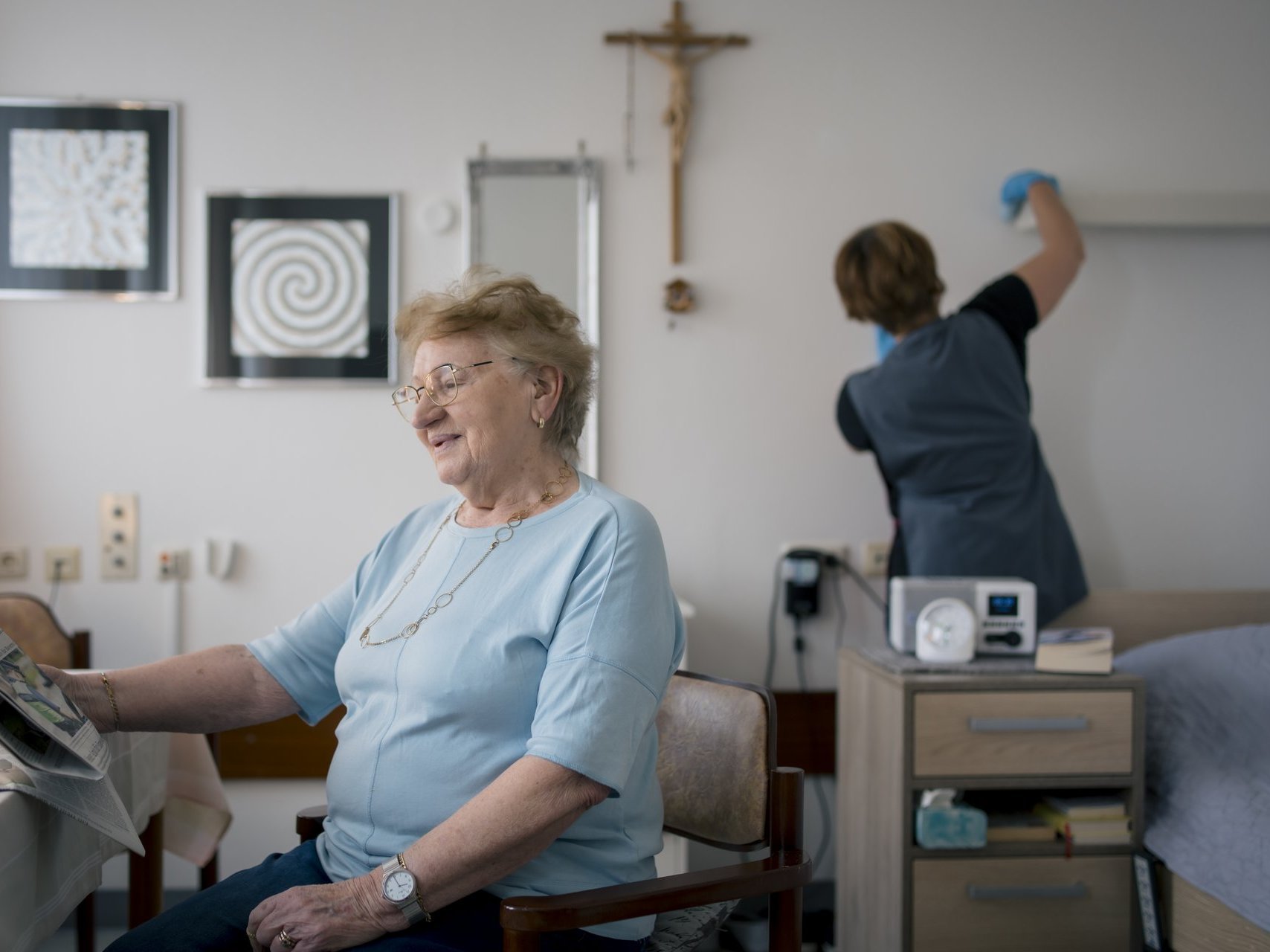 Im Vordergrund sitzt eine ältere Frau am Tisch und liest Zeitung, im Hintergrund wischt eine Frau Staub von einer Leiste an der Wand über dem Bett.