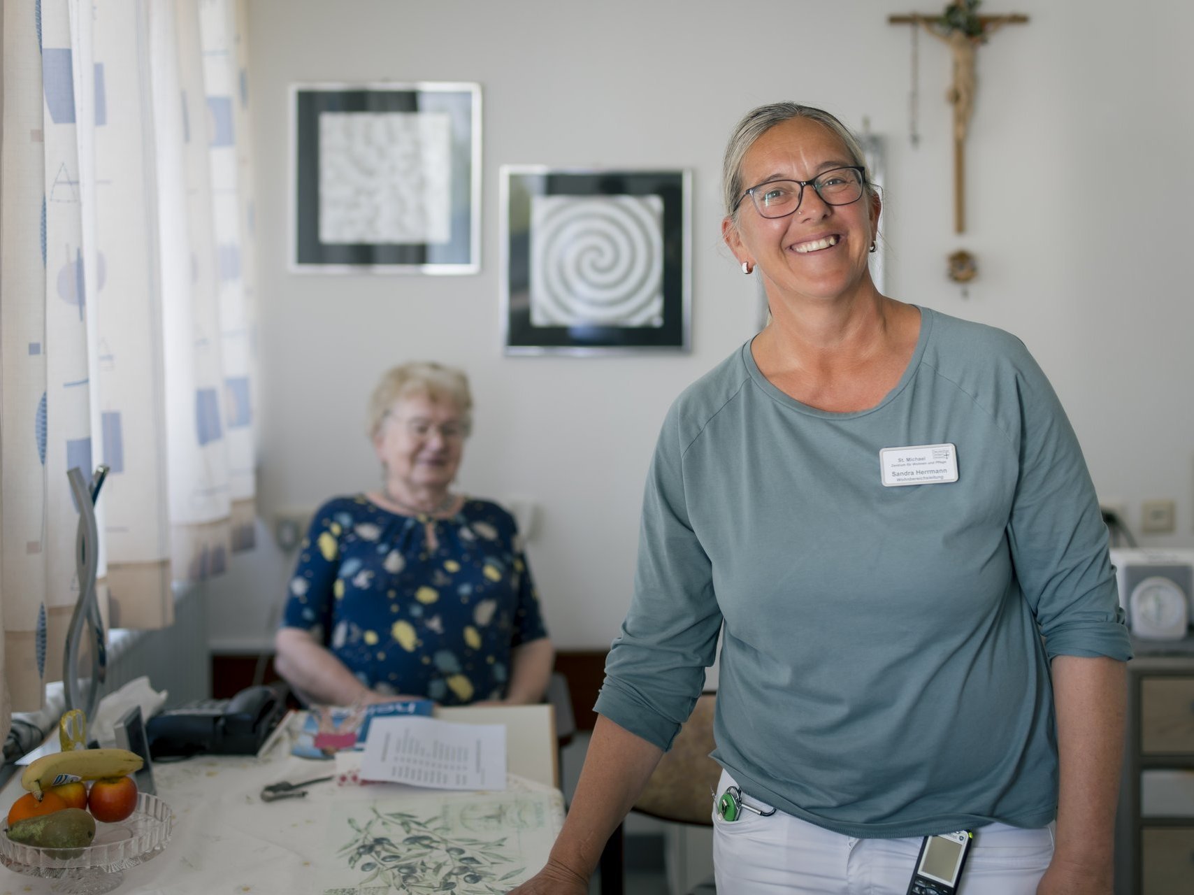 Eine ältere Dame sitzt am Tisch im Hintergrund des Fotos und sieht einer jüngeren Pflegerin nach, die sich in Richtung Kamera bewegt, beide lächeln.