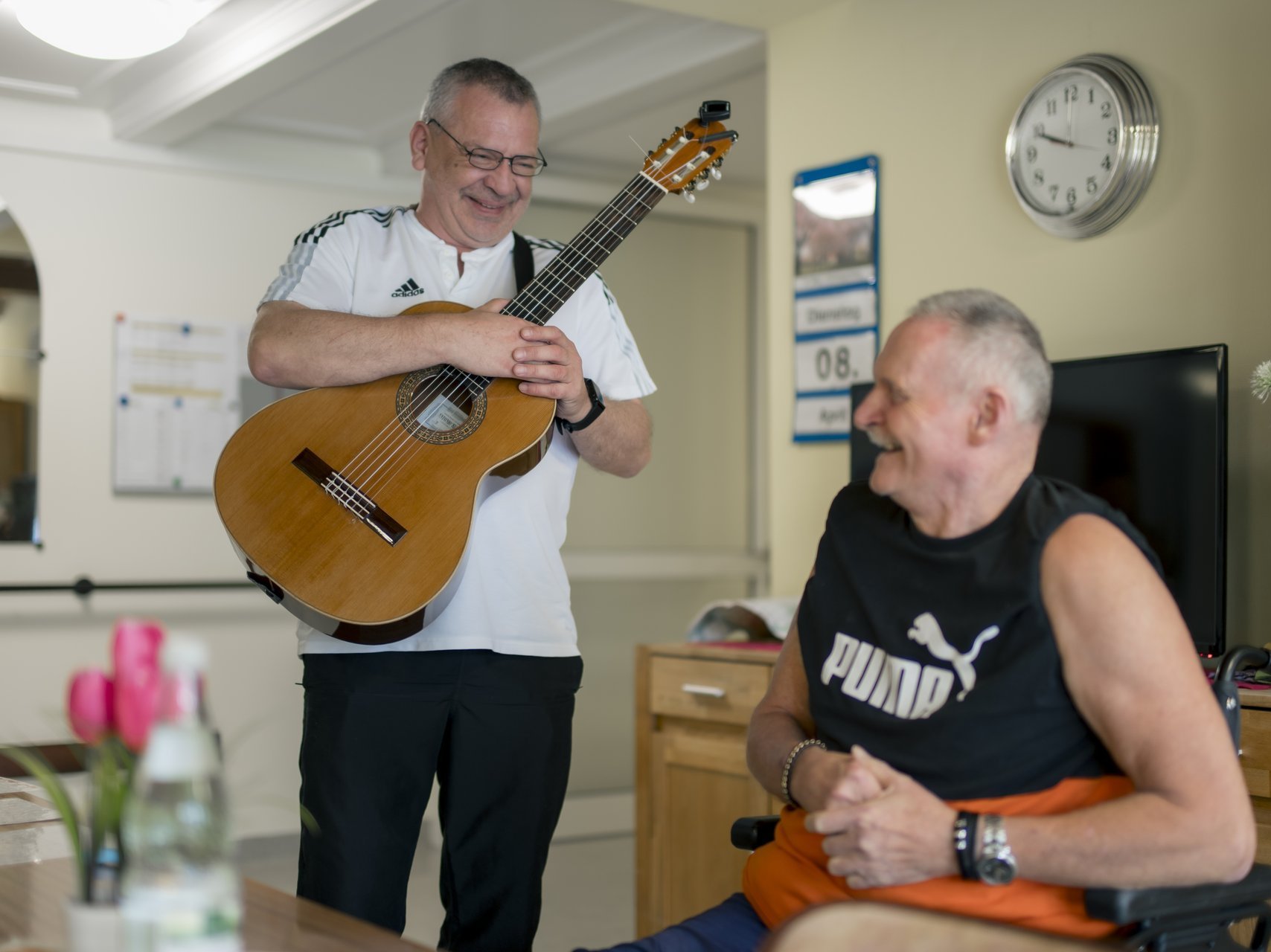Ein Herr steht mit einer Gitarre neben einem anderen, der im Rollstuhl sitzt und beide lachen.