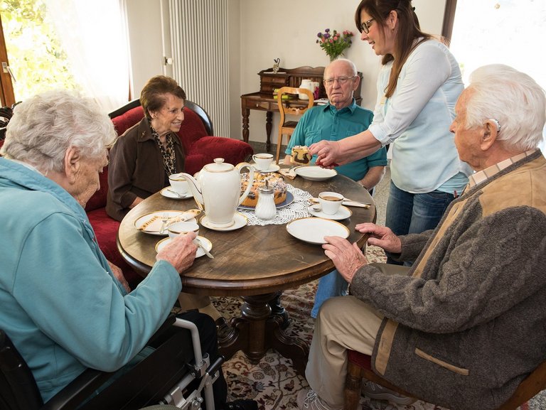 Bewohner beim Kaffetrinken an einem runden Tisch - eine Pflegerin schneidet und teilt Marmorkuchen aus