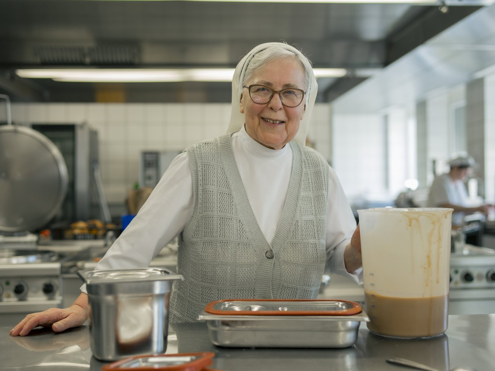Eine Ordensschwester steht an einer Arbeitsfläche in der Küche, hält einen Messbecher mit Sauce in der Hand und lächelt in die Kamera.