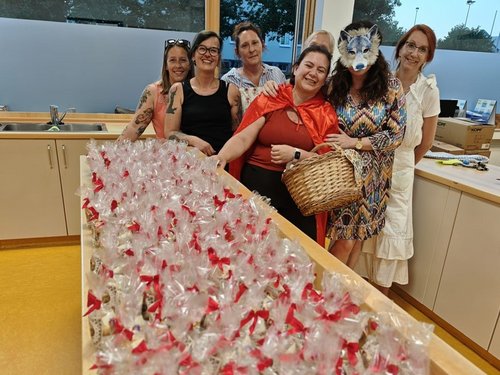 Sieben Frauen stehen in einer Küche hinter einem langen Tisch, der mit zahlreichen verpackten Leckereien und roten Schleifen gefüllt ist. Eine Frau mit rotem Umhang hält einen Korb, eine andere trägt eine Wolfsmaske.