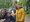 Vier fröhliche Personen posieren im Park. Eine ältere Frau sitzt im Rollstuhl, ein Mann lehnt sich von links zu ihr, und zwei Frauen stehen rechts daneben. Alle lächeln in die Kamera vor einem Hintergrund aus Bäumen und blauem Himmel.