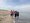 Vier ältere Menschen stehen barfuß nebeneinander am Strandrand. Zwei Frauen und ein Mann halten sich an den Armen, während die vierte Frau die Hand des Mannes hält. Im Hintergrund sind Sand, das Meer und der blaue Himmel zu sehen.