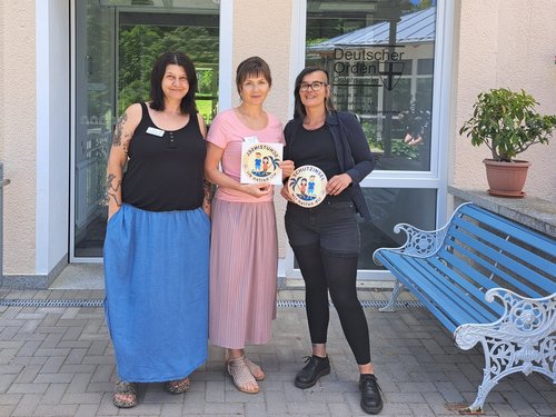 Drei Frauen stehen vor einem Gebäude und präsentieren Auszeichnungen. Die Frau in der Mitte und die Frau rechts halten jeweils ein rundes Zertifikat mit dem Logo des Deutschen Gründerpreises.