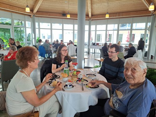 Vier Personen essen gemeinsam an einem runden Tisch in einem hellen Raum. Im Vordergrund rechts sitzt eine ältere Dame in blauem Oberteil, die in die Kamera blickt. Links und gegenüber sitzen drei jüngere Menschen. Auf dem Tisch stehen Teller mit Essen und Getränke.