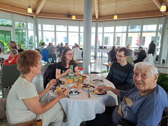 Vier Personen essen gemeinsam an einem runden Tisch in einem hellen Raum. Im Vordergrund rechts sitzt eine ältere Dame in blauem Oberteil, die in die Kamera blickt. Links und gegenüber sitzen drei jüngere Menschen. Auf dem Tisch stehen Teller mit Essen und Getränke.